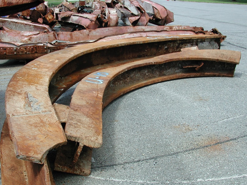 Figure 38. Heavy I-beams bent into smooth semicircles around their vertical strong axis, demonstrating orthogonal-axis deformation inconsistent with gravitational overload.
<br>
- Image by NIST.